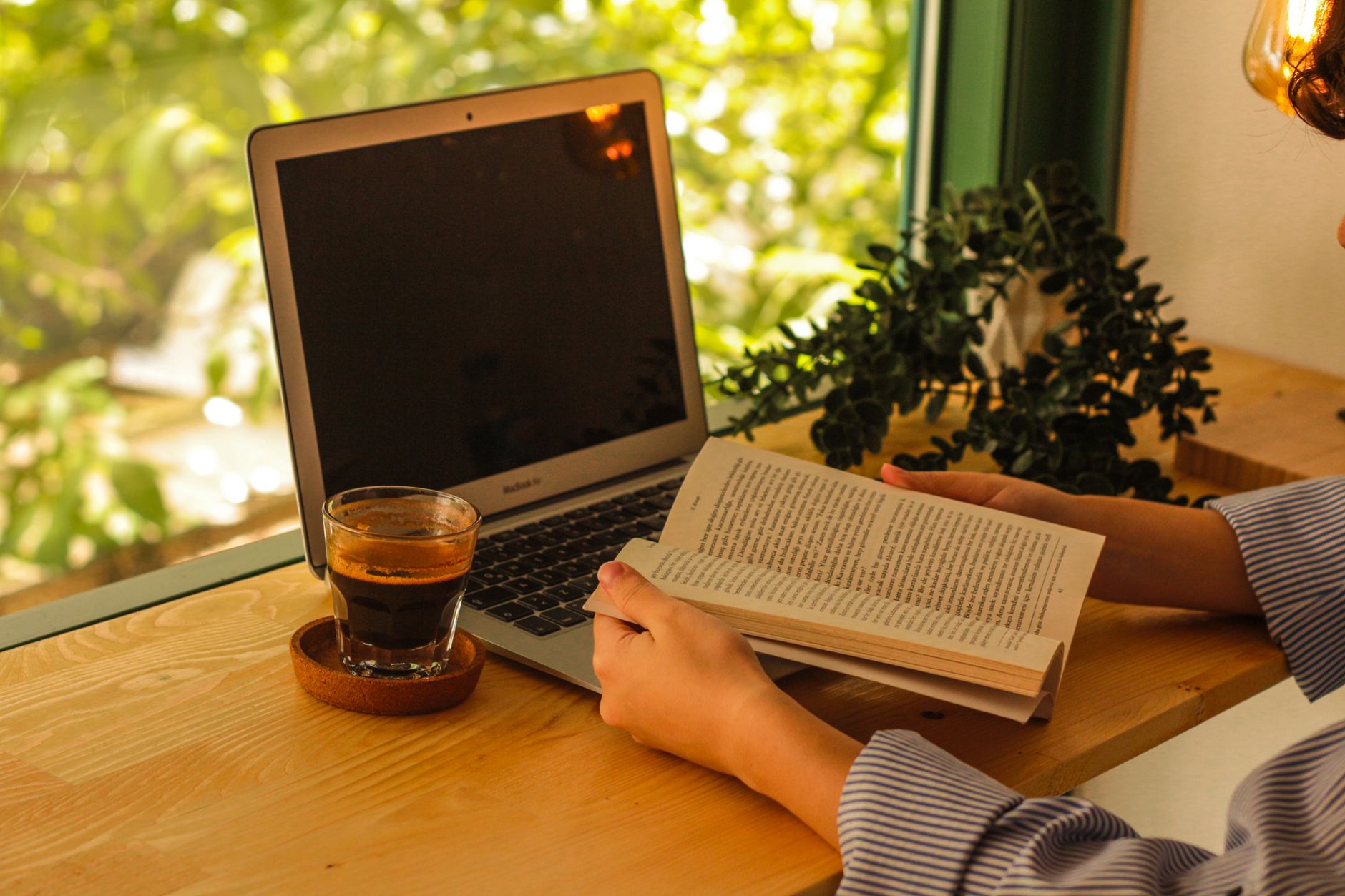 Iemand leest een boek naast een kop koffie en een laptop bij het raam